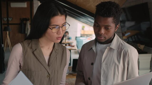 Medium close-up shot of young Asian woman with documents and African American man with laptop standing together in coworking office, looking at screen and discussing some work issue