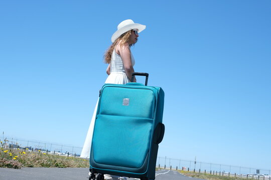 Woman Girl In White Dress Tourist With Large Blue Suitcase Goes Into Distance Adjusting Hat Against Sky Travel Runway Place For Travel Agency Advertising Text. Beautiful Developing Long Clothes