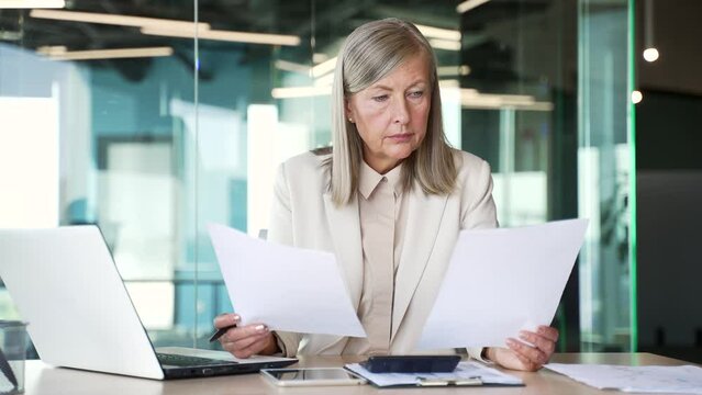 Confused puzzled senior gray haired businesswoman having difficulty with paper work sitting at workplace in business office. Frustrated accountant reviewing documents and cannot understand problem 