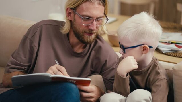 A blond man in glasses with a beard helps his little son, an albino boy in glasses with white hair, to sort out his homework assignment and read the sentence correctly. Blond man doing homework with