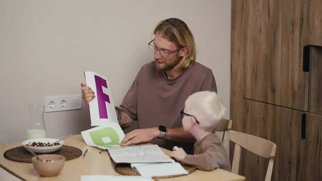A blond man in glasses with a beard shows his little albino son in blue glasses letters in English and teaches him grammar in English in preparing his homework in elementary school in the kitchen