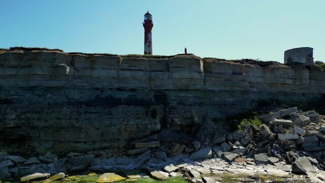 Unrecognizable person standing up high on a cliff edge. Aerial of woman standing ontop of rock cliff overlooking coastal and lighthouse in the background. Limestone in Paldiski, Estonia