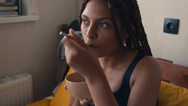 Side tilt closeup of African American lady sitting on sofa and eating whole grain cereal with blueberries for breakfast in morning and looking away in modern apartmentSide tilt closeup of African Amer