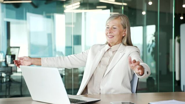 Happy senior gray haired businesswoman finished work on laptop sitting at workplace in office. Smiling satisfied mature woman puts her hands behind her head, stretches herself in a chair. Work done
