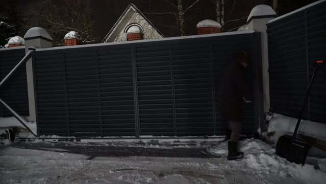 Senior man dressed in warm clothes helps with crowbar to open frozen automatic gates on snowy winter evening. Modern designer motorized automatic gates for entering courtyard in winter.