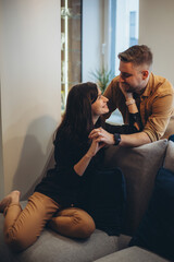 A girl in a black sweater and brown trousers is sitting on the sofa, and a guy is standing over her and hugging her. The girl holds the guy by the shirt and smiles.