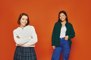 happy young multicultural women in vibrant attire standing with crossed arms on orange background