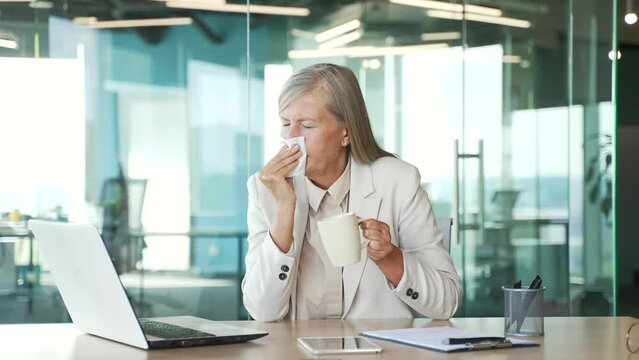 Sick senior gray haired businesswoman suffering from cold and runny nose while sitting at a workplace in a business office. Mature female wipes her nose with a handkerchief. She has a fever and flu