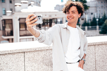 Portrait of young attractive man with curly hair hairstyle. Smiling handsome male in casual stylish clothes posing in the street at sunny day. Cheerful and happy model outdoors. Take selfie photos