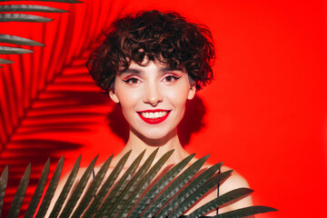 Fashion beauty portrait of young smiling brunette woman with evening stylish makeup and perfect clean skin. Sexy model with curly hair posing in studio. With red bright lips. Tropical palm leaf