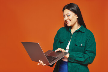 happy young asian woman in vibrant attire using laptop on orange background, remote work concept
