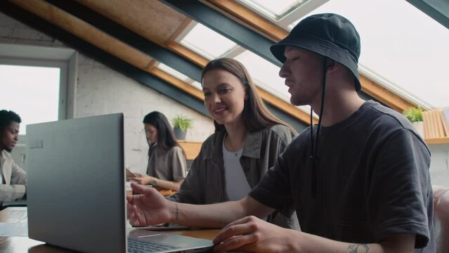 Medium shot of young Caucasian guy in t-shirt and hat sitting at table in coworking office facility, explaining his work to female acquaintance, pointing to laptop, woman listening with interest