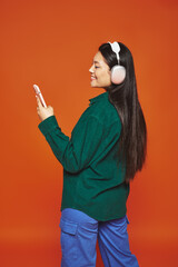 happy asian woman listening music in wireless headphones using smartphone on orange background