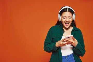 excited asian woman listening music in wireless headphones using smartphone on orange background