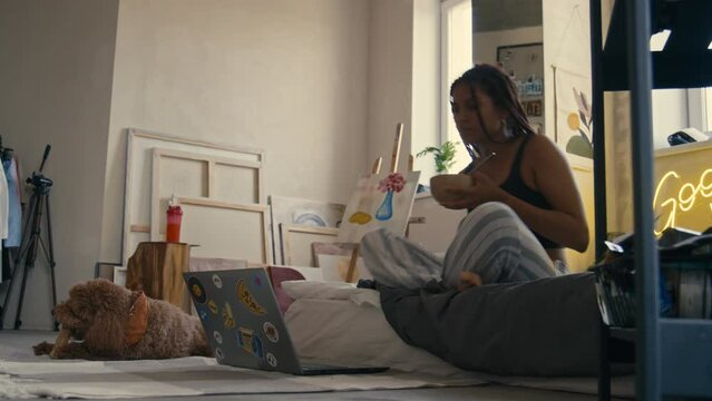 Full side low section shot of young Black girl in pajamas taking sit in bed with bowl, eating meal while watching movie or video on laptop near pet in wabi sabi loft apartmentFull side low section sho
