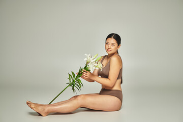 young and brunette asian woman in underwear holding flowers and sitting on grey background, lilies