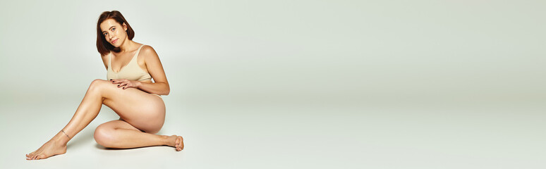 Fototapeta premium tattooed young woman with short hair posing in beige lingerie and sitting on grey background, banner