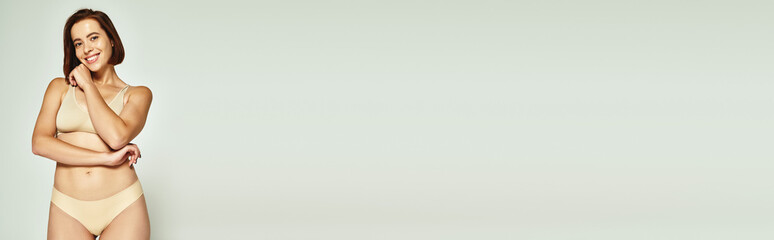 Fototapeta premium banner of young woman in underwear smiling and posing with hand near face on grey background