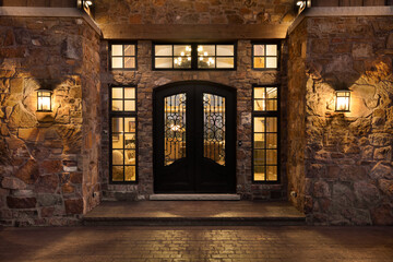A stone sided home detail with an iron door and surrounded by windows at night. Warm bulbs light...