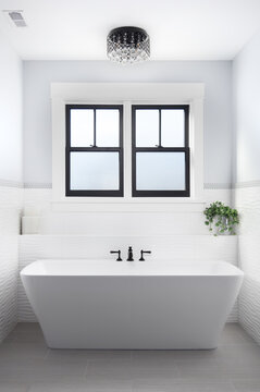 A beautiful, luxury bathroom with a light hanging above a white standalone tub and black faucet surrounded by tiles. A light with crystals hangs above.