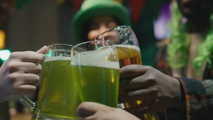 Closeup slowmo of unidentified diverse friends clanging glasses of green and craft beer in celebration of Saint Patricks day indoorsCloseup slowmo of unidentified diverse friends clanging glasses of g
