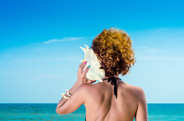 young woman with red hair with a big sea shell and colorful manicure on the background of the sea...