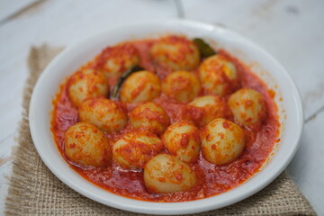 Telur Balado Tomat or Spicy Egg is traditional popular Indonesian food made from boiled egg with tomato and chili paste or sambal, served in  white plate on white wooden table. Garlic, tomato, chilli.