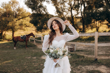 Portrait of a cute cowboy girl in a white dress with a bouquet on the background of a brown horse....