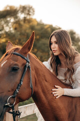 Obraz premium Portrait of a cute cowboy girl in a white dress with a bouquet on the background of a brown horse. Summer evening on a ranch farm at sunset