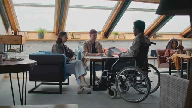 Full shot of African American man in wheelchair joining colleagues at team meeting in coworking office, greeting and submitting document draft to manager, Asian female customer sitting with smartphone