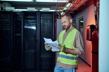 focused gray hired specialist in safety clothes looking attentively at his paperwork, data center