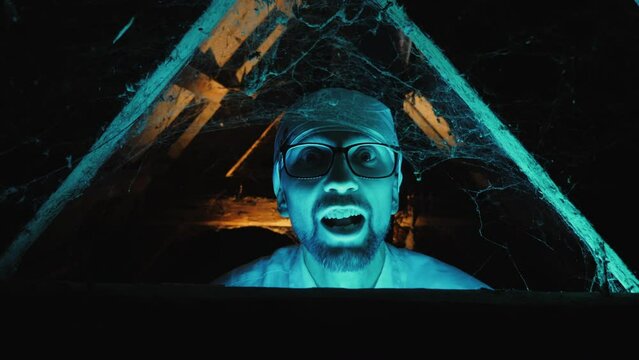 A mad scientist with glasses lit by a blue glow grimaces and screams looking at the camera, surrounded by cobwebs and wooden beams