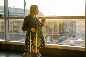 A young woman, holding a cup of coffee is ready for boarding with her suitcase.