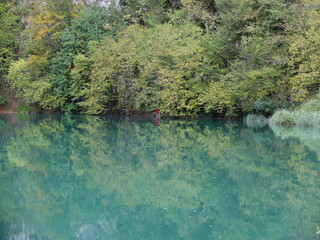 크로아티아 플리트비체 국립공원의 호수에 나뭇잎이 반영된 자연풍경으로 초록빛 호수에 연두색과 다채로운 초록빛의 나뭇잎이 비쳐 신비로운 느낌이 드며 , 비가 온 뒤 넘친 물에 잠긴 도보길을 빨간색 우비를 입고 걷고 있는 사람 1명이 포인트 입니다. 