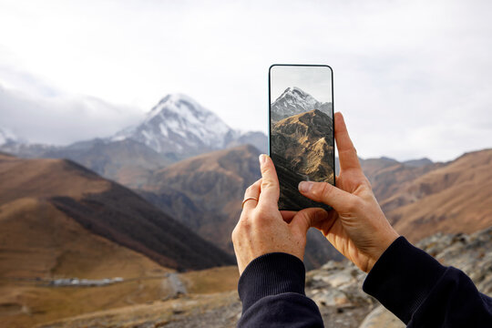 A hand holds a smartphone capturing the breathtaking view of a majestic mountain landscape.