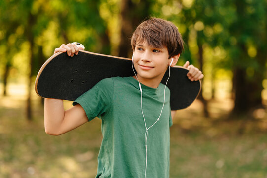 Outdoors portrait of a cool teen boy listening music in earphones and holding his skateboard.