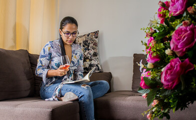 mujer joven latina usando gafas y leyendo un libro mientras sostiene una copa de vino  - obrazy, fototapety, plakaty