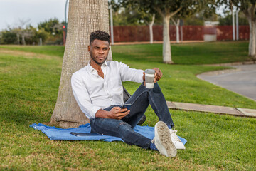 A man sits comfortably in the green grass, leaning against a tall tree, enjoying the warmth of a sunny day while holding a cell phone and a cup of coffee.