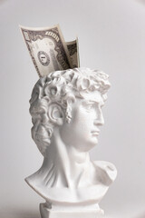 Antique male David statue's head with a wad of hundred-dollar cash banknotes isolated on a white...