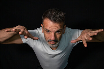 Portrait of a mature man with a gray beard, wearing braces, gesturing with his hands, wearing white t-shirt on a black background.
