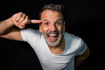 Portrait of a mature man with a gray beard, wearing braces, gesturing with his hands, wearing white t-shirt on a black background.