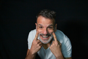Portrait of a mature man with a gray beard, wearing braces, gesturing with his hands, wearing white...