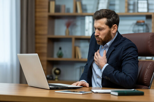 Mature businessman in a home office setup looks distressed, clutching his chest as if experiencing discomfort or pain.