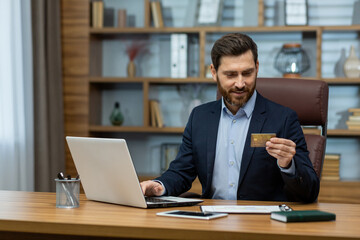 Fototapeta na wymiar Mature businessman in home office holding credit card, contemplating online payment or investment on laptop.