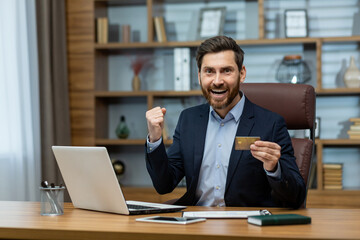 Joyful mature businessman holding a credit card and celebrating a financial triumph in a tastefully decorated home office.