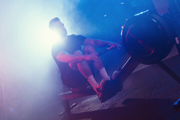 A male athlete pulls the oars on a rowing machine, his form lit by moody blue and red lights,...
