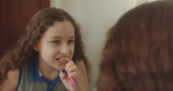 Portrait happy cute young teenage girl brushing teeth in bathroom and smiling. Children daily healthcare routine. Caucasian kid with white tooth looking at mirror isolated at home. Lifestyle. 4K