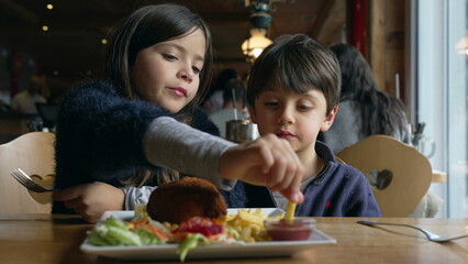 Children eating food at restaurant, siblings - small brother and sister enjoying fries and chicken meal at diner inside wooden interior