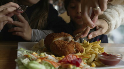 Children at restaurant about to eat cordon bleu food mealtime, closeup parent hand cutting meat, preparing for son and daughter. Little boy grabbing fry with ketchup
