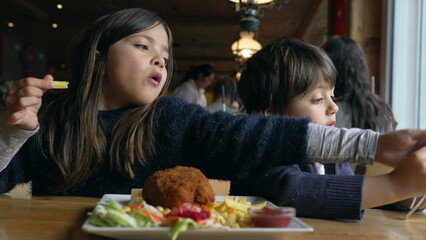 Children eating food at restaurant, siblings - small brother and sister enjoying fries and chicken meal at diner inside wooden interior